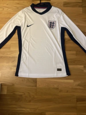 England långärmad matchtröja Nike - Säljer en oanvänd långärmad replika England fotbollströja från Nike i vitt med mörkblå detaljer längs sidorna och ärmarna. Tröjan har V-ringning, broderad Nike-logga och Englands landslagsemblem på bröstet. Perfekt för dig som älskar fotboll och vill stötta England.
