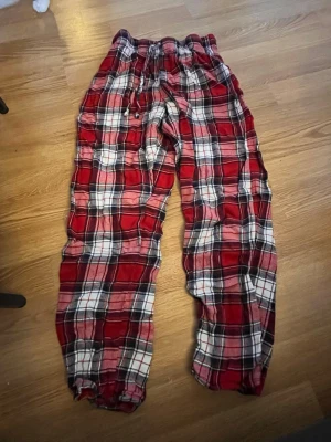 Rödrutiga mjuka loungebyxor - Säljer ett par sköna loungebyxor i rött, vitt och svart rutigt mönster. Byxorna har resår i midjan med dragsko och är tillverkade i ett mjukt, tunt tyg som passar perfekt för att chilla hemma. Avslappnad passform och långa ben.