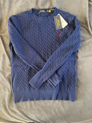 Ralph lauren tröja - Snygg blå kabelstickad tröja från Polo Ralph Lauren med rund halsringning och den klassiska röda logotypen broderad på bröstet. Tröjan har långa ärmar och ribbade muddar vid ärmslut och nederkant. Perfekt för en stilren look. OBS LITEN I STORLEKEN PASSAR NOG BÄTTRE TILL EN MEDIUM