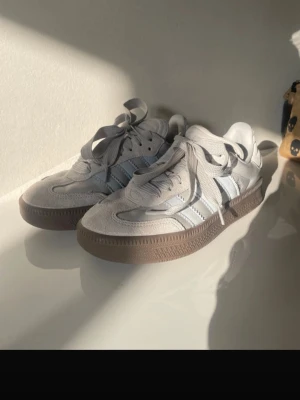 Adidas ljusgrå sneakers med mockadetaljer - Säljer ett par stilrena Adidas sneakers i ljusgrått med klassiska ränder på sidan. Skorna har ovandel i mocka och syntet, samt snörning och en brun gummisula. Perfekta för dig som gillar en clean och sportig look. Original pris 1200kr