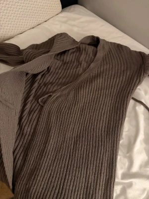 Brun ribbstickad kofta med knytband omlott klänning - Mysig brun ribbstickad kofta med långa ärmar och knytband i midjan. Koftan har en djup V-ringning och är tillverkad i ett mjukt, tjockt material som passar perfekt för kyligare dagar. Enkel och stilren design som är lätt att matcha.