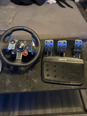 Logitech ratt och pedalset PS4/PC - Säljer ett Logitech ratt- och pedalset med racingratt i svart läder och blå detaljer, samt tre metallpedaler på en robust svart platta. Perfekt för racing på PS4 och PC, med realistisk känsla och många knappar för kontroll. Allt funkar bra de ända problemet är att de saknas en kabel till ratten 