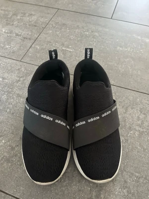 Svarta slip-on sneakers från Adidas - Säljer ett par svarta slip-on sneakers från Adidas med breda elastiska band över foten där Adidas-loggan syns. Ovandelen är i mesh-liknande material och sulan är vit. Skorna har dragflikar bak för enkel påtagning och en modern, sportig look.