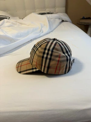 Burberry klassisk rutig keps - Säljer en ikonisk keps från Burberry i klassiskt beige rutigt mönster med svarta, vita och röda linjer. Kepsen har böjd skärm och justerbar rem med metallknappar bak. Tillverkad i bomull och perfekt för dig som vill ha en tidlös accessoar.