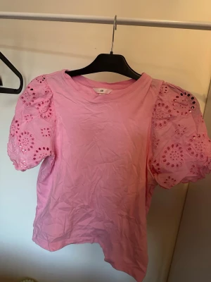 Rosa topp med puffärm från H&M - Söt rosa topp från H&M med korta puffärmar i broderad spets och rund halsringning. Ärmarna har fina hålbroderier som ger en romantisk vibe. Perfekt att styla med jeans eller kjol för en trendig look.