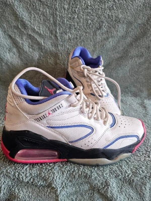 Vita Air Jordan sneakers med blå detaljer - Säljer ett par vita Air Jordan sneakers med blå och rosa detaljer. Skorna har svart sula med rosa inslag och klassisk Jumpman-logga på plösen. Ovandelen är i skinn med perforerade partier och snörning. Insidan är fodrad i blått tyg för extra komfort.