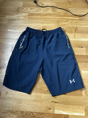 Blå träningsshorts från Under Armour - Säljer ett par mörkblå träningsshorts från Under Armour med elastisk midja och dragkedjor på fickorna. Shortsen har logotypen på ena benet och är tillverkade i lätt syntetmaterial som passar perfekt för sport och aktivitet.