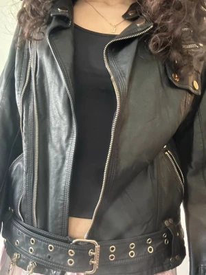 Svart skinnjacka med bälte och detaljer - Cool svart skinnjacka med flera dragkedjor, knappar och ett brett bälte med metallhål. Jackan har klassisk bikerstil och snygga detaljer som ger en edgy look. Perfekt för dig som vill ha en statement piece i garderoben.