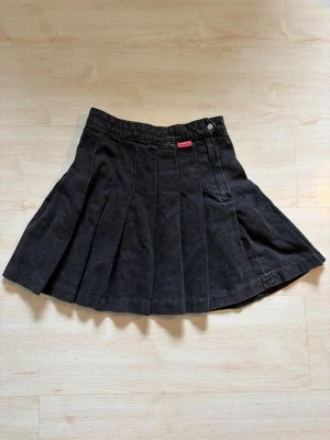 Svart kortkjol - Säljer en svart plisserad kortkjol med klassisk design och knappdetalj i midjan. Perfekt för att skapa en cool och trendig look.