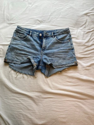 Blå denim shorts med fransig kant - Säljer ett par klassiska blå denim shorts med fransig kant och råa avslut. De har knapp och  dragkedja framtill samt en avslappnad passform. Perfekta för varma dagar och en chill look.