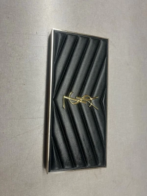 YSL Couture Colour Clutch ögonskugga - Lyxig ögonskuggspalett från Yves Saint Laurent med svart quiltad design och guldfärgad YSL-logga på locket. Paletten har en elegant och exklusiv look med metalliska detaljer och kommer i en guldig förpackning. Perfekt för dig som älskar high-end smink och snygg design.