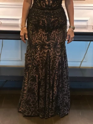 Svart långklänning med spetsdetaljer - Elegant svart långklänning med transparenta spetsdetaljer och vackert blommönster. Klänningen har markerad midja och figurnära passform upptill, samt en lätt utsvängd kjol. Perfekt för dig som vill sticka ut på bal eller fest.