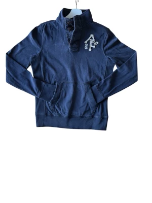 Mörkblå hoodie från Abercrombie & Fitch - Snygg mörkblå hoodie från Abercrombie & Fitch med hög krage och knappar. Stor broderad logga på bröstet och känguruficka framtill. Tillverkad i mjuk bomull, perfekt för chill dagar. Klassisk och stilren design som passar till det mesta.