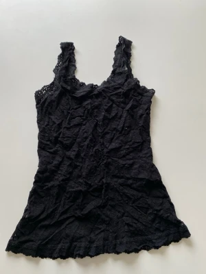 Svart spetslinne med breda axelband - Ett svart spetslinne med breda axelband och dekorativa spetsdetaljer längs kanterna. Materialet är mjukt och lätt, perfekt för fest klädsel. 