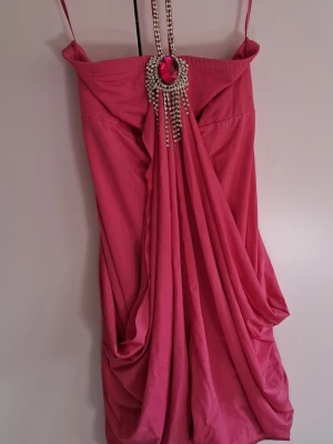 Rosa kortklänning med strassdetalj - Snygg rosa kortklänning med tunna axelband och draperad front. Klänningen har en stor rosa sten och strasskedjor som hänger ner från halsen, vilket ger en glamorös look. Perfekt för dig som vill sticka ut och känna dig extra fin. Är några stenar som lossatdärför jag säljer den billigt