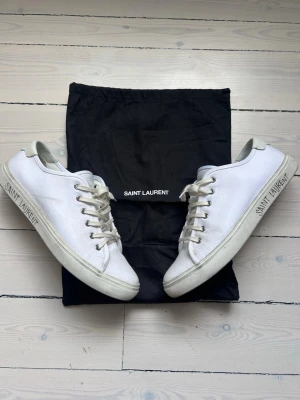 Vita Saint Laurent canvas sneakers - Snygga vita sneakers från Saint Laurent i canvas med klassisk låg profil och vita snören. Märkets logga syns på sidan och innersulan. Skorna har en stilren design och kommer med svart dustbag.