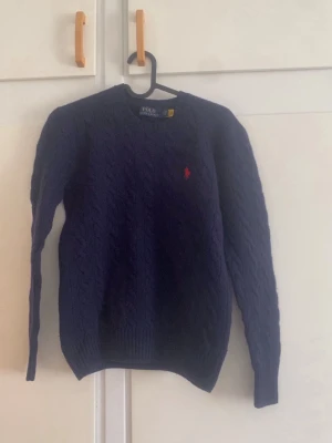 Mörkblå kabelstickad tröja Polo Ralph Lauren - Mörkblå kabelstickad tröja från Polo Ralph Lauren med rund hals och lång ärm. Tröjan har ett klassiskt rött broderat logomärke på bröstet och ribbstickade muddar. Perfekt för dig som gillar stilrena och tidlösa plagg.