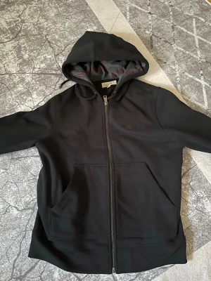 Svart hoodie med dragkedja Burberry - Säljer en svart hoodie från Burberry med dragkedja och huva. Insidan av huvan har klassiskt rutmönster i grått och rött. Hoodien har två stora fickor framtill och är tillverkad i mjukt material som känns skönt mot huden. Perfekt för dig som vill ha en stilren och exklusiv look.