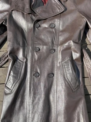Brun lång skinnjacka med dubbelknäppning - Snygg brun skinnjacka med dubbelknäppning och bred krage. Jackan har två snedställda fickor framtill och ett rött innerfoder. Perfekt för dig som vill ha en cool och tidlös look. Storlek 54 enligt etiketten. Dock behöver ena fickan lite kärlek den behöver sys. 