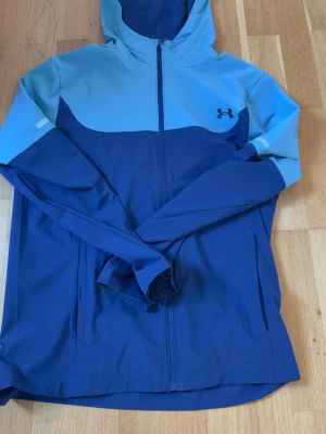 Blå vindjacka från Under Armour - Snygg vindjacka från Under Armour i två nyanser av blått med huva och hel dragkedja. Jackan har en sportig design med logotyp på bröstet och är tillverkad i ett lätt material som passar perfekt för utomhusaktiviteter. Praktiska fickor och bekväm passform.