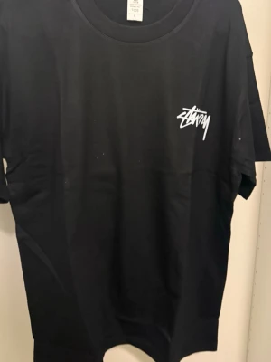 Svart Stüssy t-shirt med tärningar - Cool svart t-shirt från Stüssy med vit logga på bröstet och stort tryck med två tärningar på ryggen. Klassisk rund hals och korta ärmar, perfekt för dig som gillar streetwear och grafiska prints. Materialet är mjuk bomull som känns skönt mot huden.