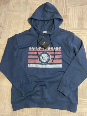 Blå hoodie från Saint Laurent - Cool blå hoodie från Saint Laurent med huva och dragsnören. Tryck på bröstet med text och emblem i rött, vitt och blått. Mjuk bomullskänsla och klassisk passform. Perfekt för dig som vill ha en snygg och avslappnad look.