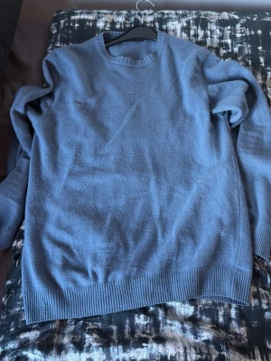 Blå stickad tröja med rund hals, oscar jacobsson - En enkel och stilren blå stickad tröja med rund hals och ribbade muddar vid ärmslut och nederkant. Perfekt för lager-på-lager och passar till jeans eller chinos. Materialet är mjukt och behagligt, vilket gör den skön att bära under kyliga dagar.(ull) 