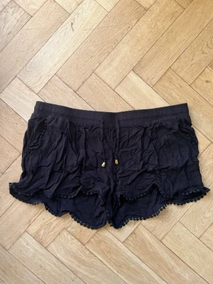Svarta shorts med snörning och spets - Säljer ett par svarta shorts med elastisk midja och snörning med guldfärgade detaljer. Shortsen har dekorativ spetskant och små bollar längs nederkanten. De är tillverkade i mjukt bomullsmaterial och har en loose passform.