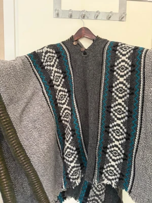 Grå poncho med mönster  - Poncho i grått med breda ärmar och dekorativa mönster i vitt, svart och turkos längs fram och bak. Har två knappar vid halsen och gröna detaljer vid ärmslut. Stickad i tjockt material, perfekt för lager-på-lager och en cool look.