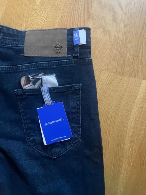 Mörkblå jeans från Jacob Cohën - Snygga mörkblå jeans från Jacob Cohën med klassisk femficksmodell och knäppning med silverfärgade knappar. Jeansen har en exklusiv känsla med detaljer som en beige patch bak och medföljande näsduk i fickan. Perfekta för dig som gillar stilrena och lyxiga jeans.