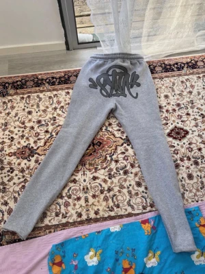 Grå mjukisbyxor med tryck - Säljer ett par grå mjukisbyxor från Syna world med coolt svart graffiti-tryck på baksidan och logga på framsidan. Byxorna har dragsko i midjan och sidofickor. Perfekta för chill och streetwear-stil.