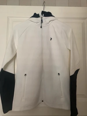 Vit hoodie med svart detalj från Peak Performance - Snygg vit hoodie från Peak Performance med svarta detaljer på huva och ärmar. Hel dragkedja framtill, två fickor med dragkedja och diskret logotyp på bröstet. Perfekt för dig som gillar stilren design och sportig känsla.