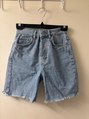 Högmidjade ljusblå jeansshorts - Säljer ett par högmidjade ljusblå jeansshorts från bik bok med råa, fransiga benslut och klassiska femficksdetaljer. Shortsen har en knappar framtill samt en läderpatch med logga bak. Perfekta för en avslappnad stil.