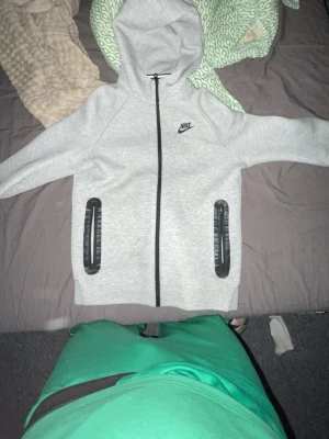 Grå zip hoodie från Nike - Snygg grå hoodie från Nike med dragkedja framtill och svart Nike-logga på bröstet. Två fickor med svarta detaljer och huva. Perfekt för chill dagar eller när du vill ha en sportig look. Tillverkad i mjukt sweatshirtmaterial.