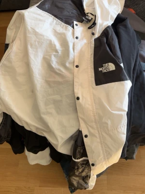Svartvit vindjacka från The North Face - Snygg svartvit vindjacka från The North Face med dragkedja och tryckknappar. Jackan har en luva med snörning och logga på bröstet. Tillverkad i lätt och tåligt material, perfekt för blåsiga dagar.  Obs dragkedjan funkar inte men den finns knappar så man kan knäppa hela jackan ändå