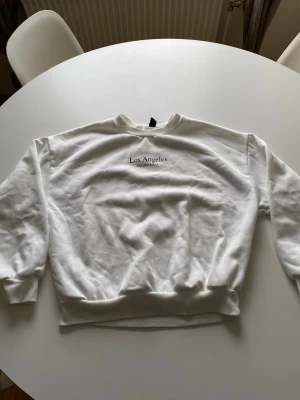 Vit sweatshirt med Los Angeles-tryck - Vit sweatshirt med rund hals och tryck 'Los Angeles California' på bröstet. Tröjan har ribbade muddar vid ärmslut och nederkant, samt en avslappnad passform. Perfekt för dig som gillar enkel och stilren streetwear.