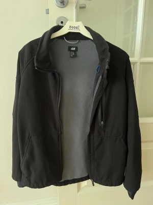 Svart vindjacka från H&M - Svart vindjacka från H&M med mjukt grått fleecefoder. Jackan har dragkedja framtill, hög krage och två sidofickor samt en bröstficka med dragkedja. Perfekt för blåsiga dagar och enkel att matcha med olika outfits.