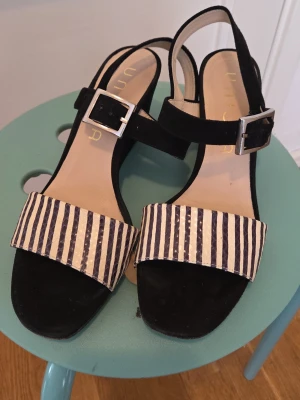 Randiga kilklackssandaler från Unisa - Snygga sandaler från Unisa med svart kilklack och breda remmar framtill i svart och vitt randigt mönster. Justerbar rem med silverfärgat spänne runt ankeln. Skorna har öppen tå och är tillverkade i mocka och textil.