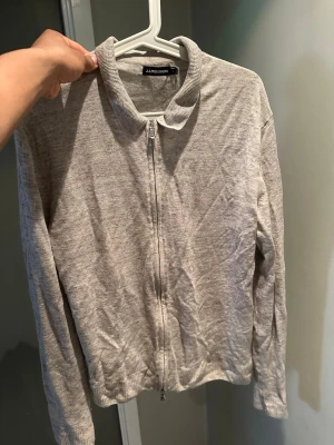 Ljusgrå ziptröja från j Lindberg  - Ljusgrå ziptröja från J Lindberg med långärmad design och ribbad krage. I storlek M.  Tröjan har en enkel och clean look, perfekt för att slänga på sig när det är lite kyligt. Materialet känns mjukt och lätt, och dragkedjan framtill gör den enkel att ta på och av.