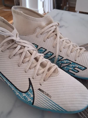 Nike Mercurial vita fotbollsskor - Säljer ett par vita Nike Mercurial fotbollsskor med högt skaft och snörning. Skorna har blå och svarta detaljer, inklusive en stor Nike-logga och mönstrade inslag på sidan. Tillverkade i syntetmaterial för bra grepp och känsla på planen.