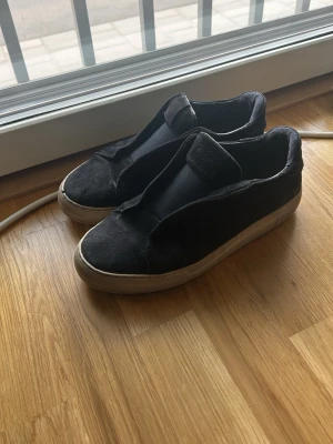 Svarta slip-on sneakers från Arigato - Lite stora i storlken. Jag har storlek 42,5. Svarta sneakers från Arigato med slip-on design och breda elastiska band över foten. Skorna har en vit, robust gummisula och är tillverkade i ett mjukt material som ger en clean och modern look. Perfekt för dig som gillar stilrena och enkla sneakers.