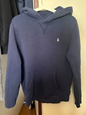 Mörkblå hoodie från Polo Ralph Lauren - Säljer en mörkblå hoodie från Polo Ralph Lauren med klassisk huva och känguruficka. Diskret logga broderad på bröstet. Perfekt för dig som gillar stilrena och bekväma plagg. Materialet är mjukt och känns riktigt skönt mot huden. Size 18/20 men motsvarar s.