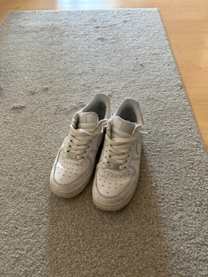 Nike Air Force 1 vita sneakers - Klassiska vita Nike Air Force 1 sneakers med låg profil och perforerad tå för extra ventilation. Skorna har en robust gummisula med cirkulärt mönster och ikonisk Swoosh på sidan. Perfekt för dig som vill ha en tidlös och clean look. Kan gå ner i pris vid snabb affär.