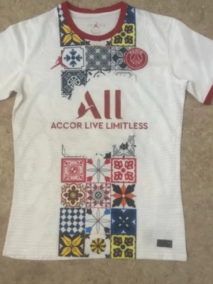 PSG matchtröja med färgglada mönster - Säljer en vit PSG matchtröja från Nike med röda detaljer vid hals och ärmslut. Tröjan har färgglada patchwork-mönster på framsidan och klubbens logga på bröstet. Materialet är lätt och andas, perfekt för träning eller match.