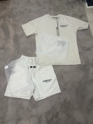 Ljusgrå Essentials T-shirt Fear of God  - Snygg ljusgrå T-shirt från Essentials Fear of God med tryck på bröstet. Klassisk rund hals och korta ärmar. Tillverkad i mjuk bomull för en skön och avslappnad look. Perfekt till streetwear-stilen.