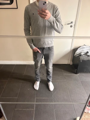 Grå stickad tröja från Polo Ralph Lauren - Jag är 165. Snygg grå stickad tröja från Polo Ralph Lauren med V-ringning och diskret logga broderad på bröstet. Tröjan har långa ärmar och ribbade muddar vid ärmslut och nederkant. Perfekt för lager-på-lager och enkel att matcha med jeans.