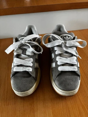 Grå Adidas Campus 00 - Snygga grå sneakers från Adidas med klassiska vita ränder på sidorna och cremevit sula. Skorna har en låg profil och är gjorda i mocka med vita skosnören och gummisula. Perfekta för en avslappnad streetstyle-look.