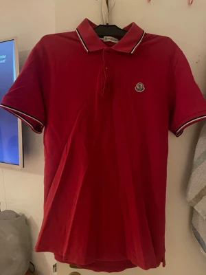 Röd pikétröja från Moncler - Snygg röd pikétröja från Moncler med klassisk krage och knäppning framtill. Tröjan har vita och svarta ränder på krage och ärmslut samt Moncler-logga på bröstet. Säljer min brors tröja. Tröjan ska vara äkta.