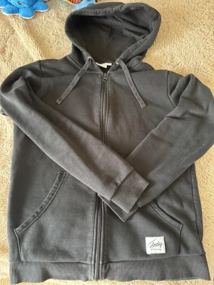 Svart hoodie från Andy by Frank Dandy - Svart hoodie med dragkedja från Andy by Frank Dandy. Klassisk modell med huva och dragsnören, två fickor framtill och ribbade muddar vid ärmar och nederkant. Tillverkad i mjukt bomullsmaterial som känns skönt mot huden.