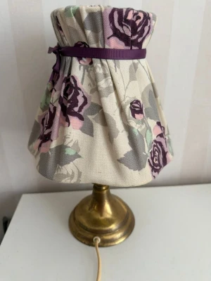Bordslampa med blommig lampskärm - Säljer en bordslampa med mässingsfärgad fot och tygskärm i vitt med lila och rosa rosor samt gröna blad. Skärmen har ett dekorativt lila band upptill. Perfekt som mysbelysning i sovrum eller vardagsrum.